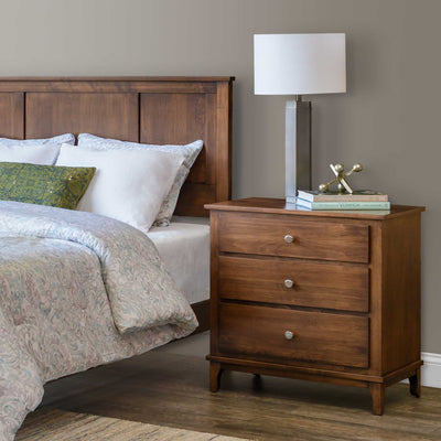 Three drawer nightstand in medium brown wood with round knobs beside a paneled headboard and lamp