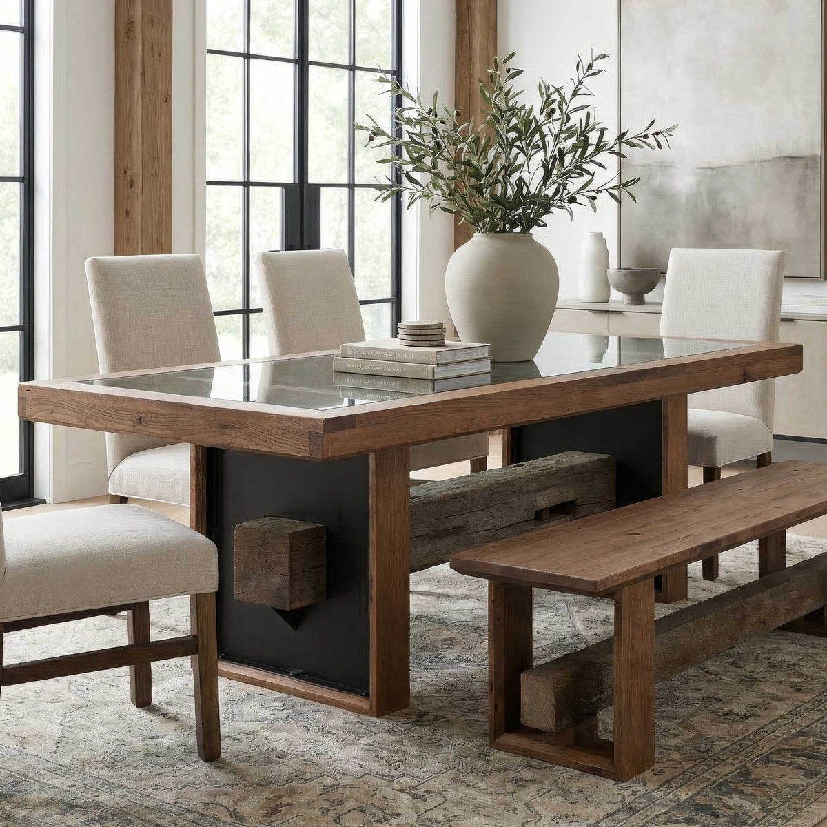 A rustic rectangular dining table with a glass top, thick wooden legs, and a black metal side panel, shown in a bright dining room with four cream upholstered chairs, a matching bench, a large vase of greenery, and books on top.