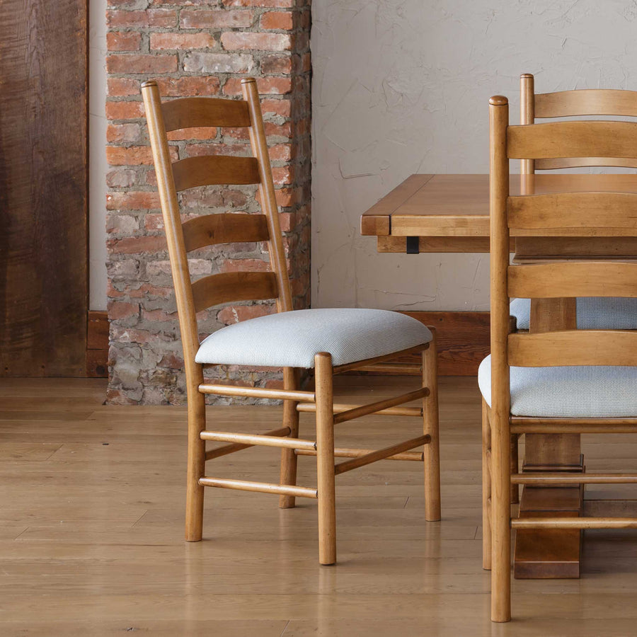 Two light brown ladder back dining chairs with pale blue upholstered seats at a plank dining table by an exposed brick wall