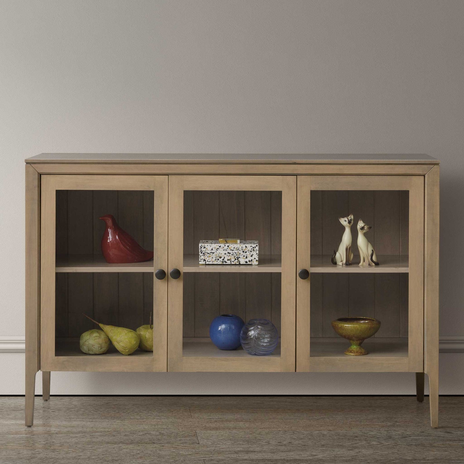 Three door glass front TV cabinet in a light driftwood finish, styled with pottery and fruit on its interior shelves