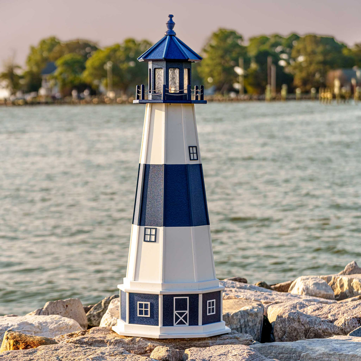 Navy and white banded lighthouse with blue lantern room on a white base, standing on rocks beside calm bay water