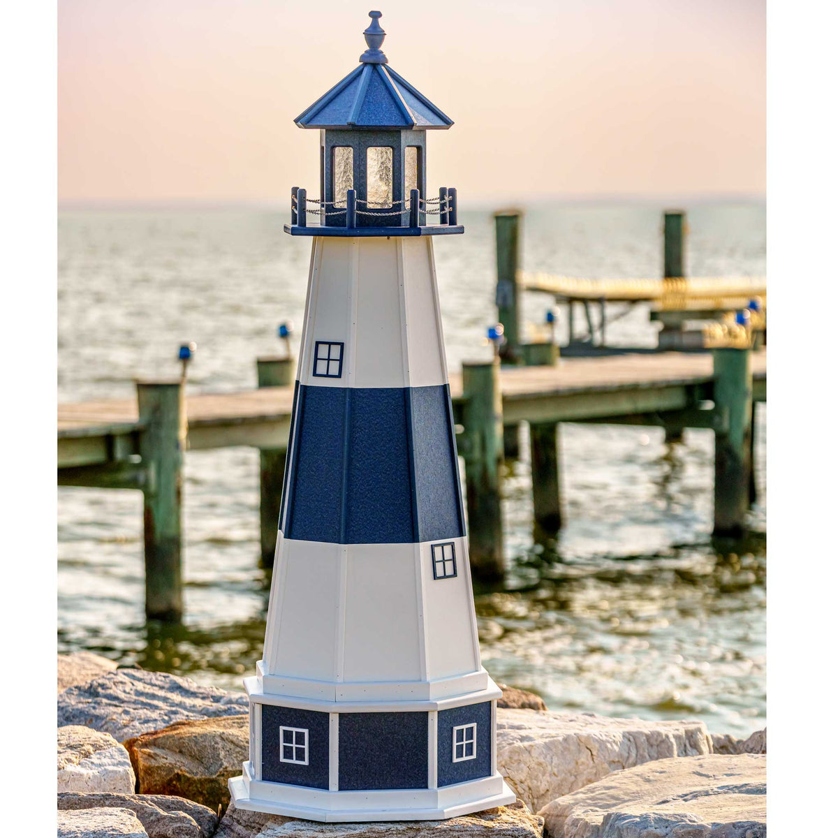 Blue and white striped lighthouse with navy paneled base on a rocky shore, wooden dock in the background