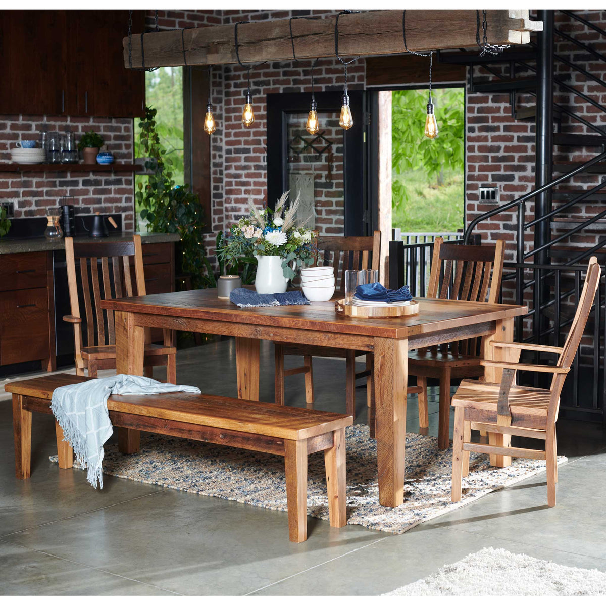 Reclaimed barnwood dining table with six slat back chairs and a bench in a brick loft kitchen with pendant lights