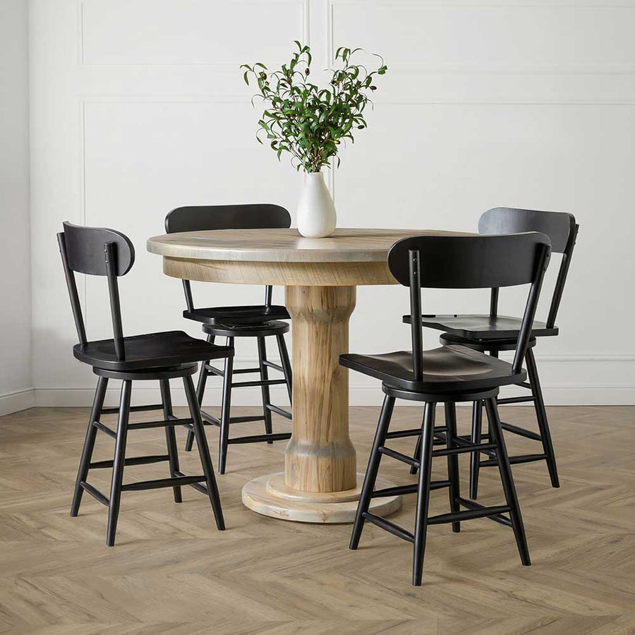 Light natural round pedestal table with four black swivel counter stools on a herringbone wood floor
