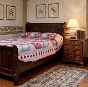Dark cherry sleigh bed with paneled scroll headboard and footboard beside a four-drawer nightstand and pink quilt