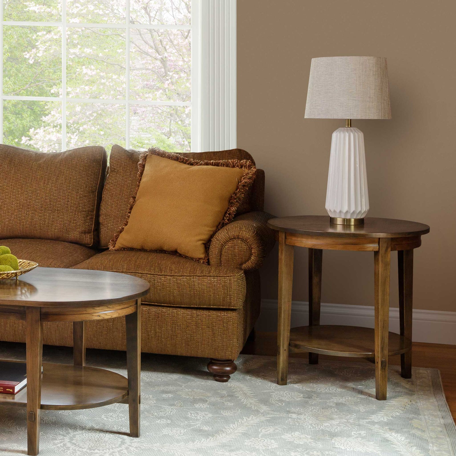 Round wood end table with a lower display shelf holding a fluted lamp beside a brown sofa in a sunlit living room