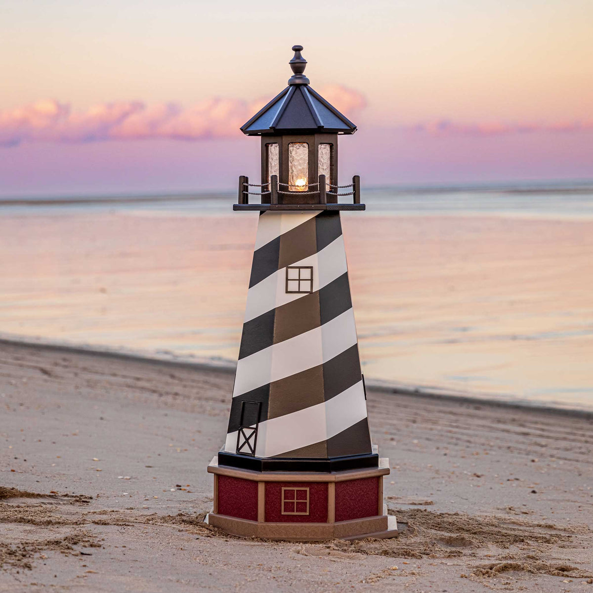 Black and white spiral striped lighthouse with lit lantern room on a red brick look base, standing on a beach at sunset
