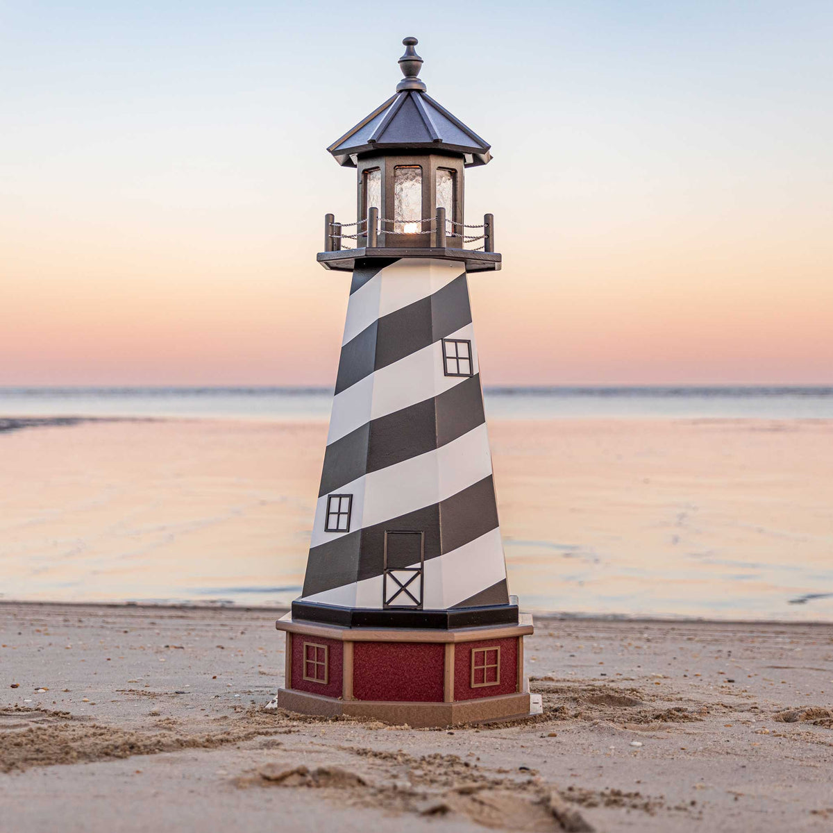 Black and white striped lighthouse with lit lantern room and red brick look base, photographed on sand at dusk