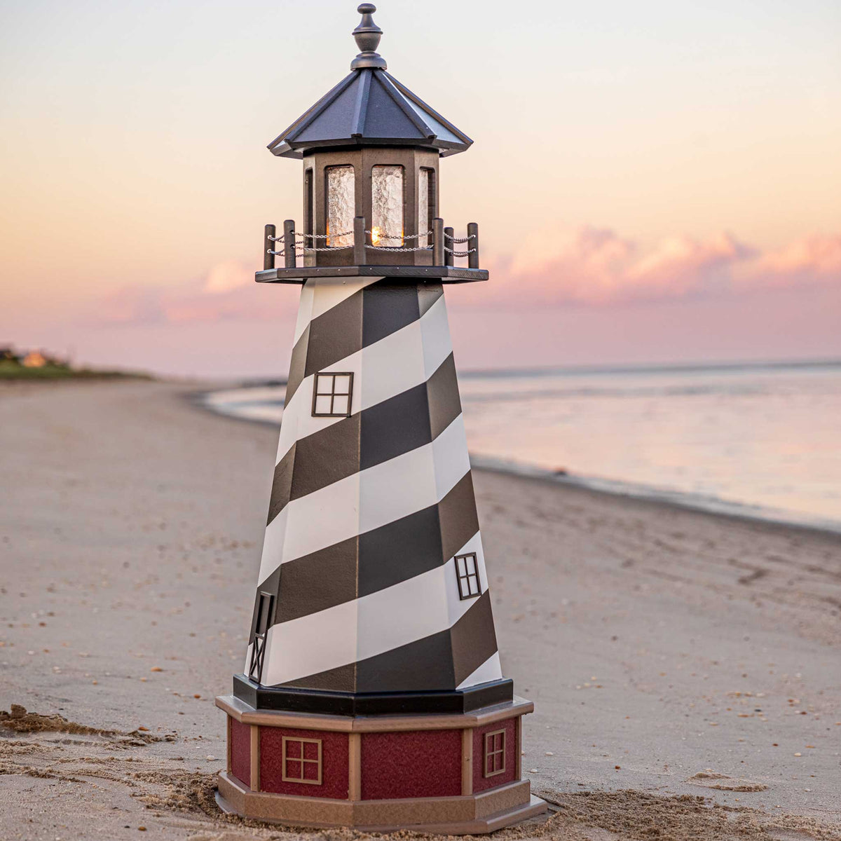 Spiral striped lighthouse with lit lantern room and red brick base standing on a sandy beach at pink sunset