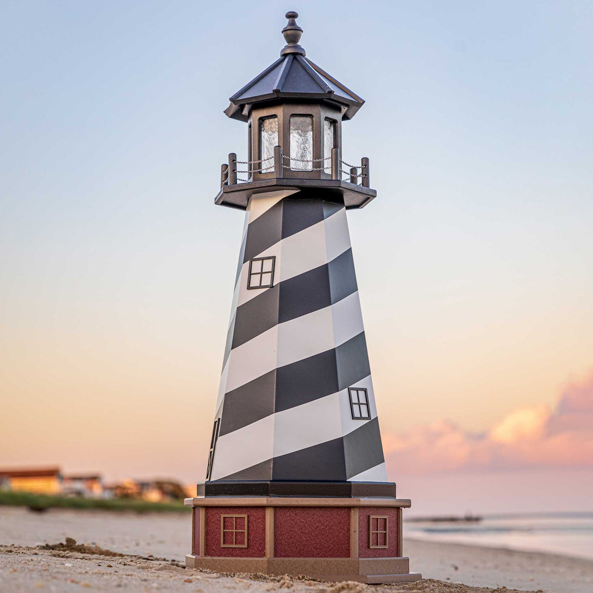 Close view of a black and white striped lighthouse with red base on a beach, ocean and dunes in the background