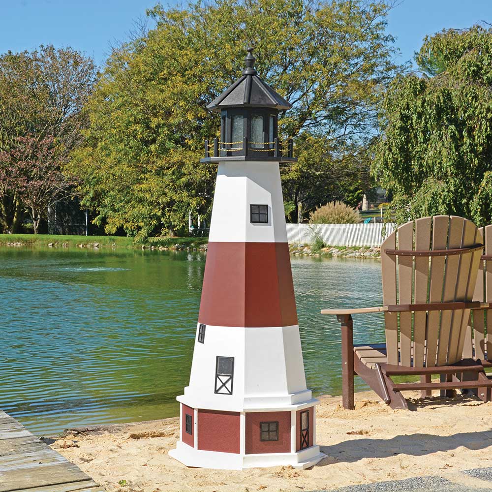 White lighthouse with a brick red band and black lantern room, set on sand by a pond next to an Adirondack chair