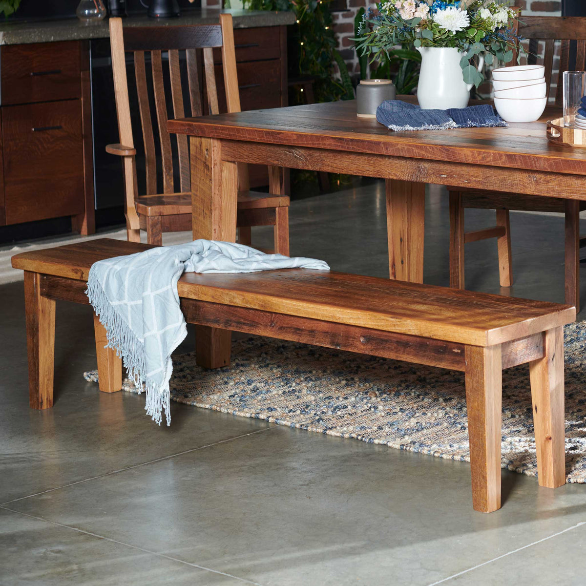 Reclaimed wood dining bench with a light throw blanket draped over the plank seat, tucked beside a rustic table