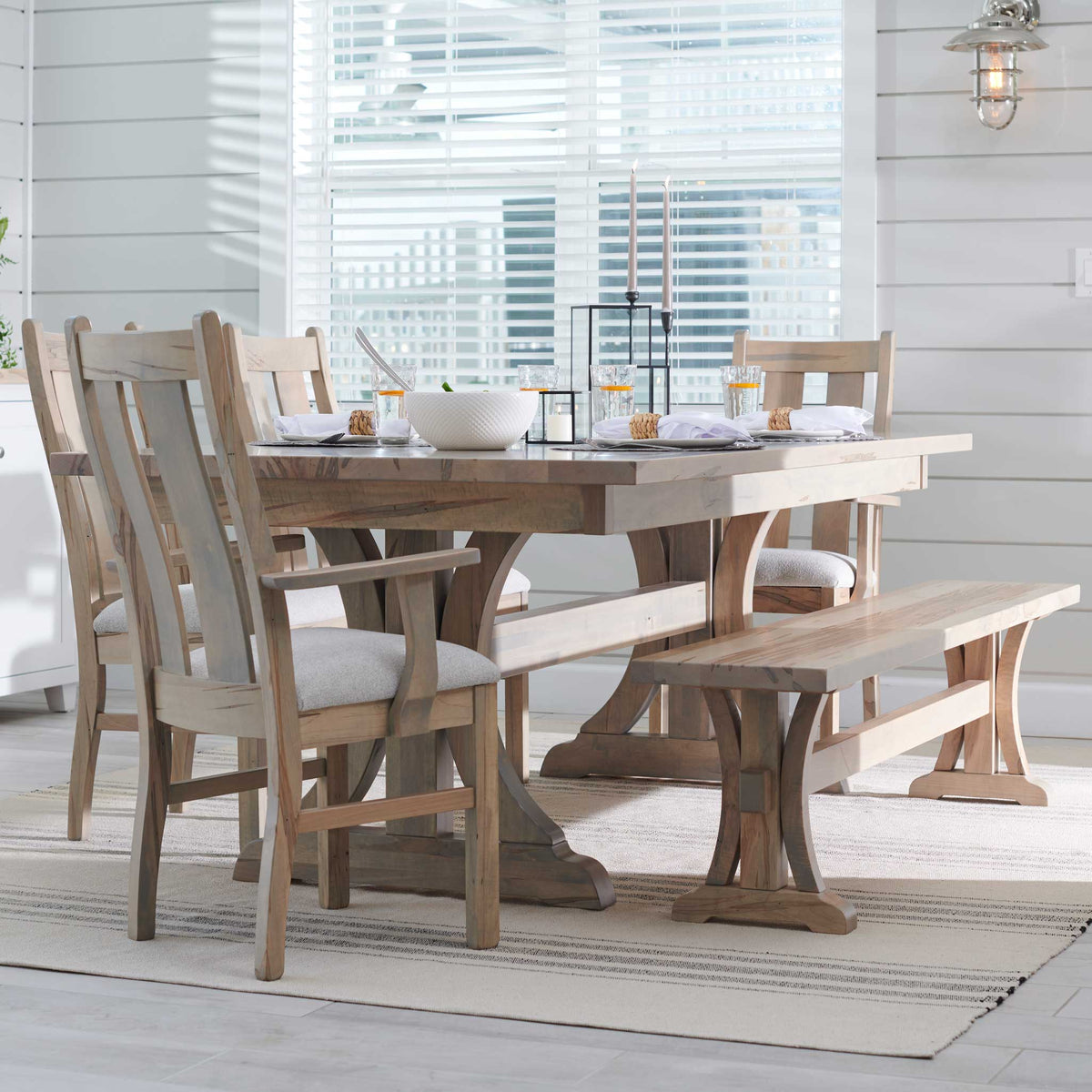Coastal dining room with a whitewashed maple trestle table set for a meal, slat-back chairs and a matching bench