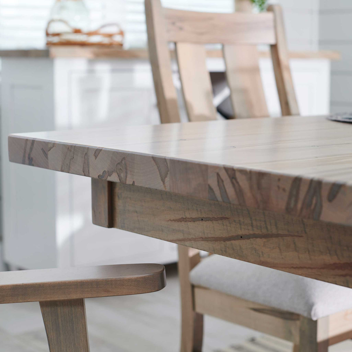 Close-up of the maple dining table's thick square edge and figured grain with a slat-back chair behind it