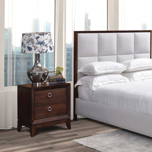 Two drawer nightstand in dark cherry with silver ring pulls, beside a gray upholstered panel bed and city window