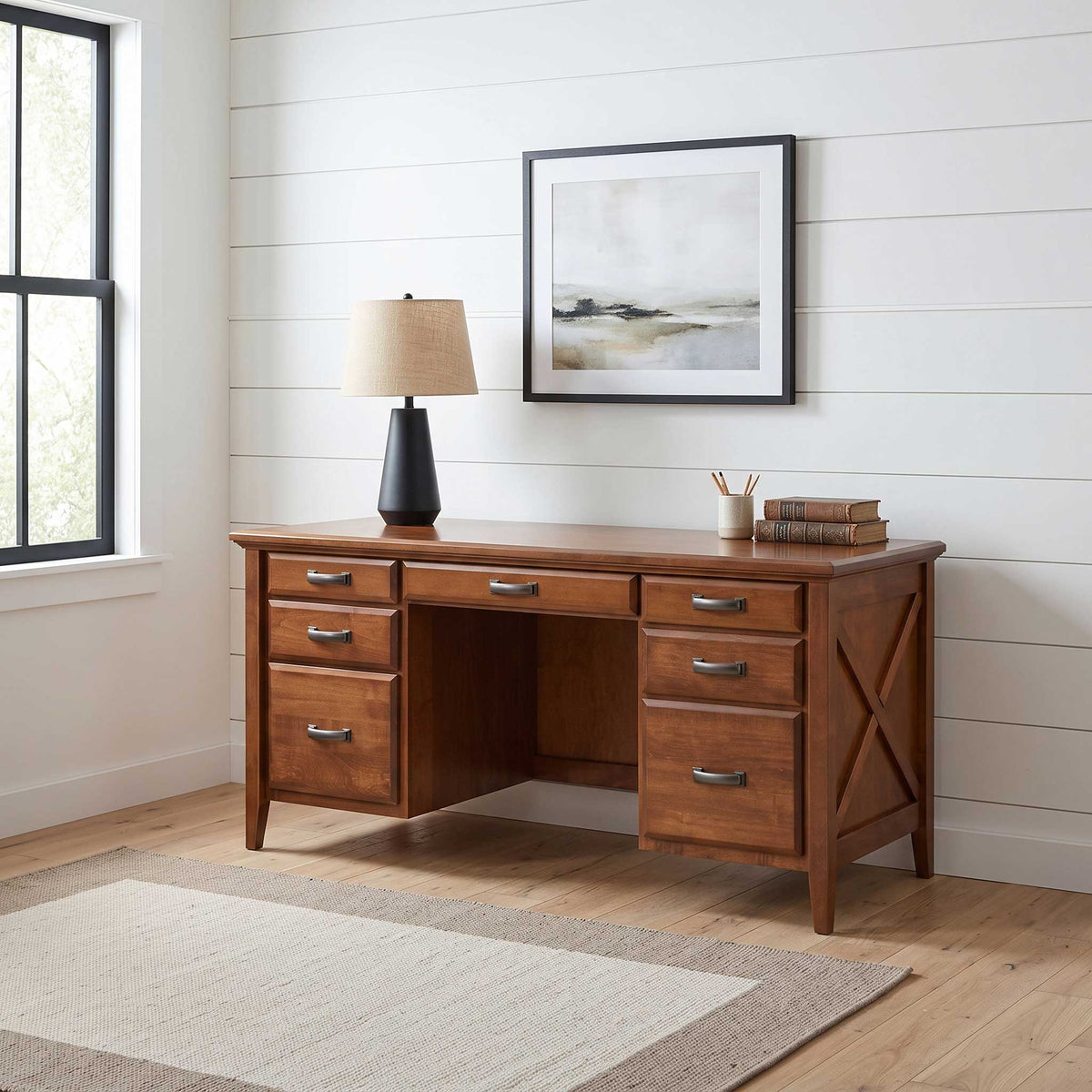 A mid-century modern writing desk in warm medium brown wood, styled in a bright home office with a black base lamp, framed wall art, and a light wood floor.
