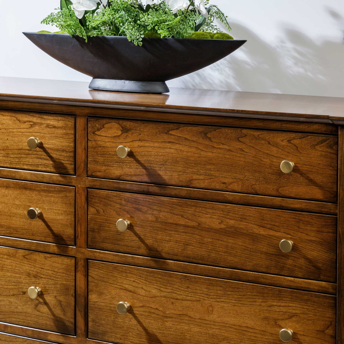 Cherry double dresser drawer fronts with brass knobs, topped by a black boat-shaped bowl of white flowers and ferns