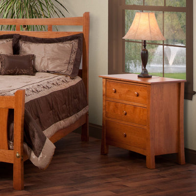 Cherry three-drawer bedside chest with round knobs and flared legs beside a bed, holding a table lamp