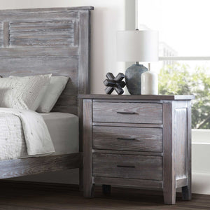 Rustic gray oak nightstand with three drawers styled beside a louvered panel bed with lamp and decor on top