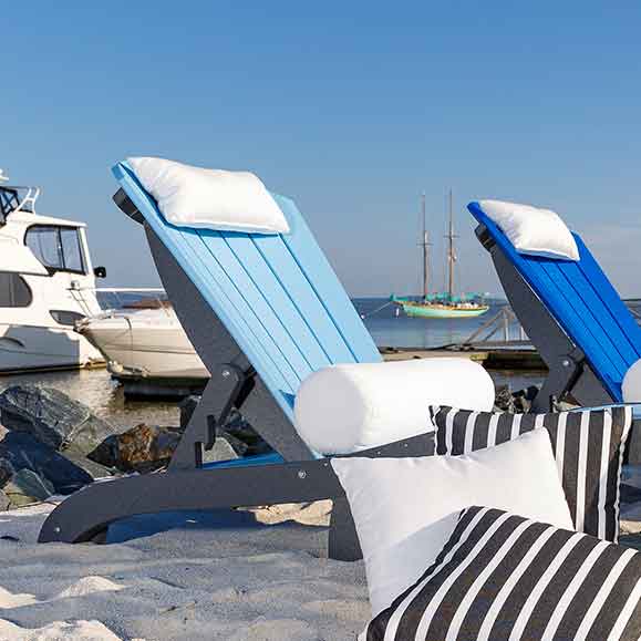 White bolster and pillows on blue poly chaise lounges on the sand beside a marina
