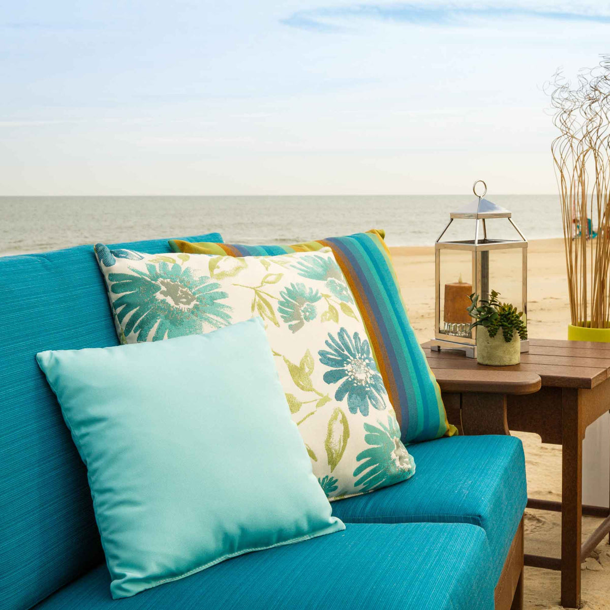 Aqua, teal floral and striped outdoor pillows on a turquoise cushioned poly sofa at the beach with a lantern nearby