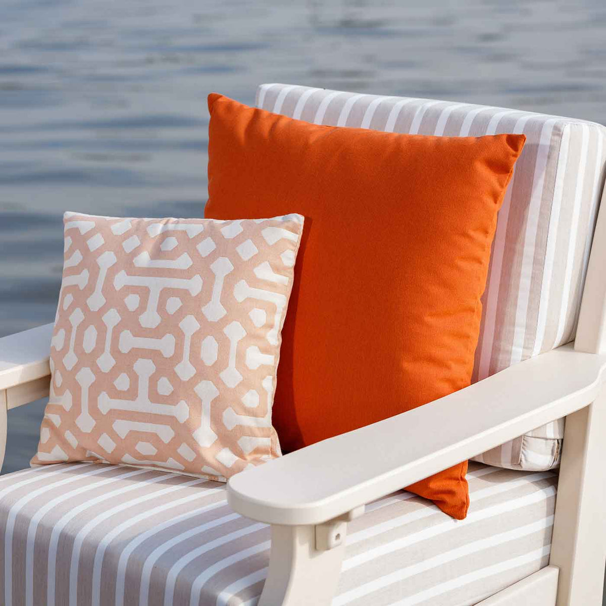 Orange solid and blush trellis print Sunbrella pillows resting on a white poly patio chair with striped cushions by the water