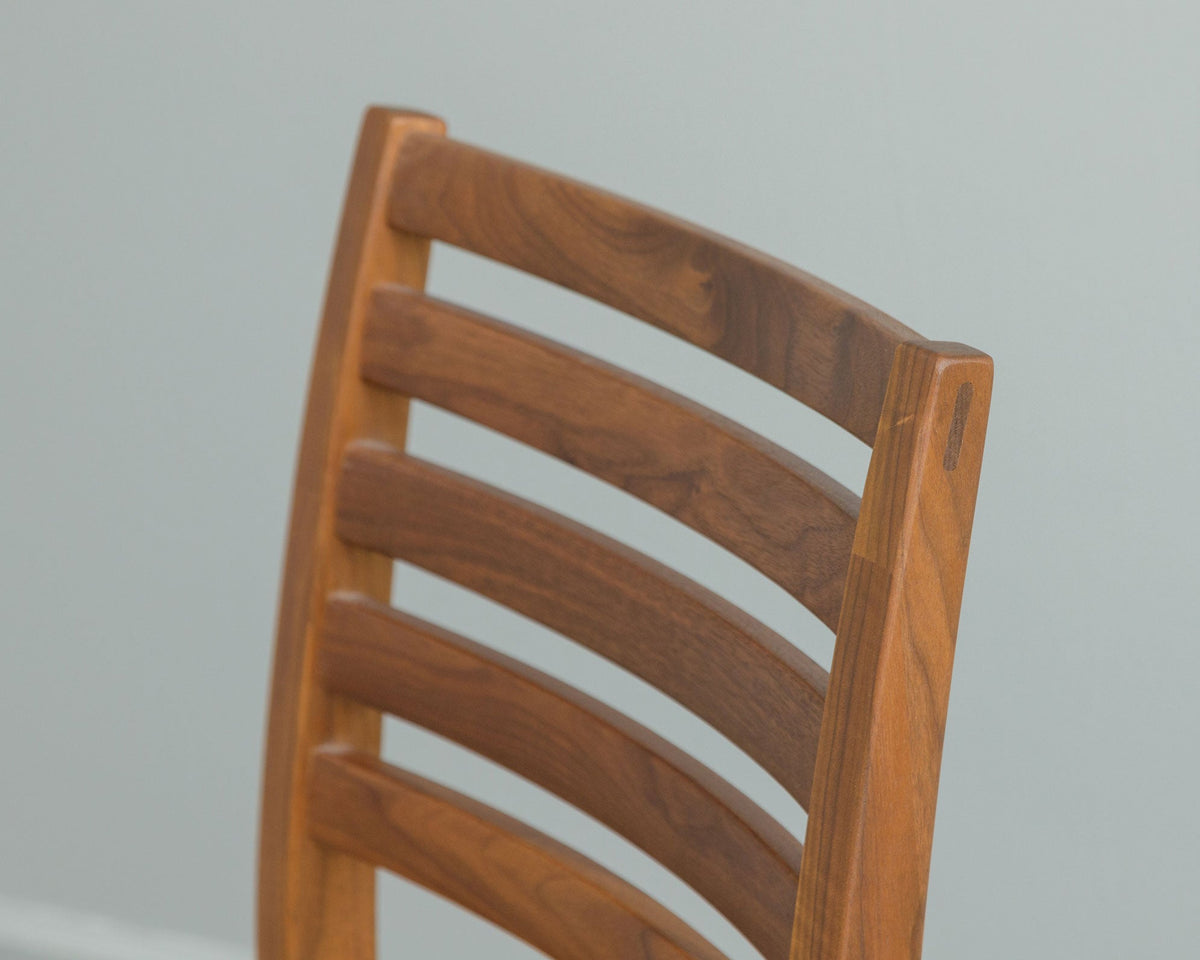 Detail of the curved slat back on a walnut stool with a visible pegged joint at the rear post