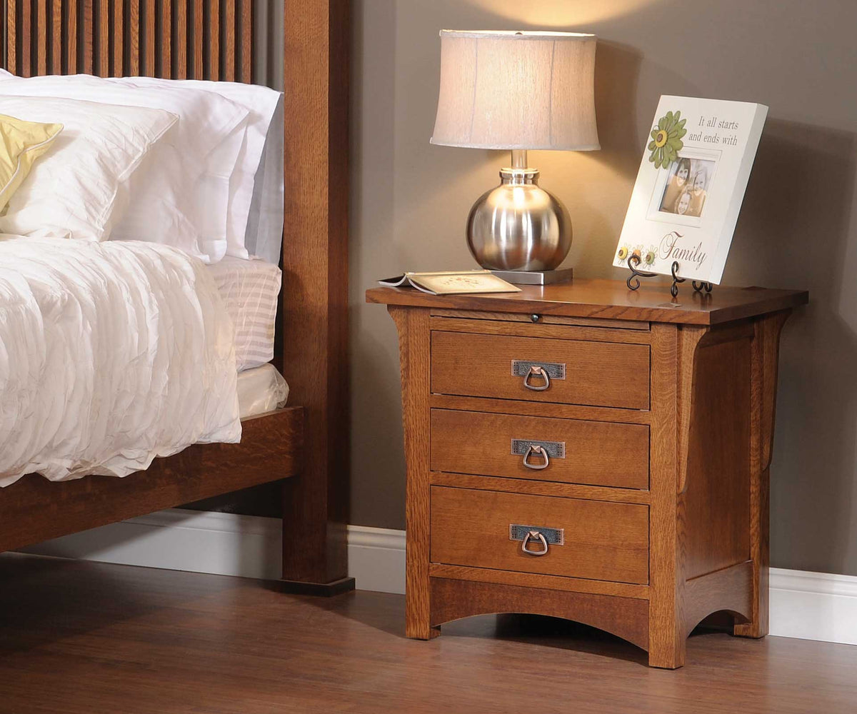 Oak three-drawer nightstand with pullout tray beside a bed, hammered ring pulls, curved sides and arched base rail