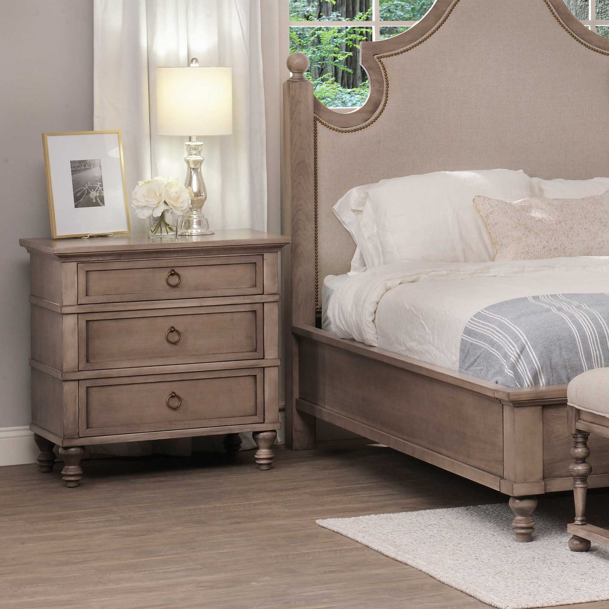 Weathered taupe three-drawer nightstand with ring pulls and turned feet beside an upholstered bed and lamp