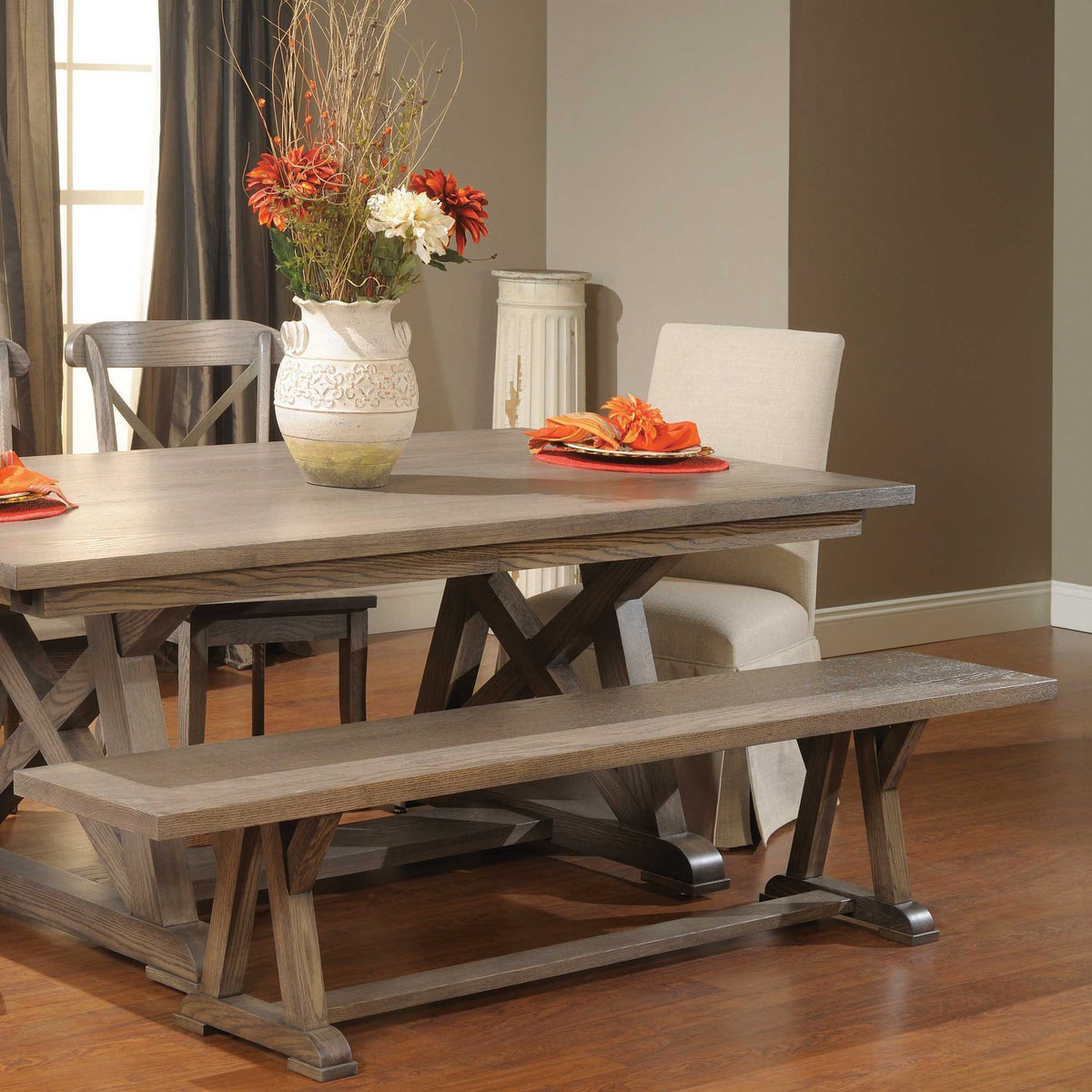 Gray oak trestle dining table with bench, cross back chairs and a floral centerpiece in a dining room