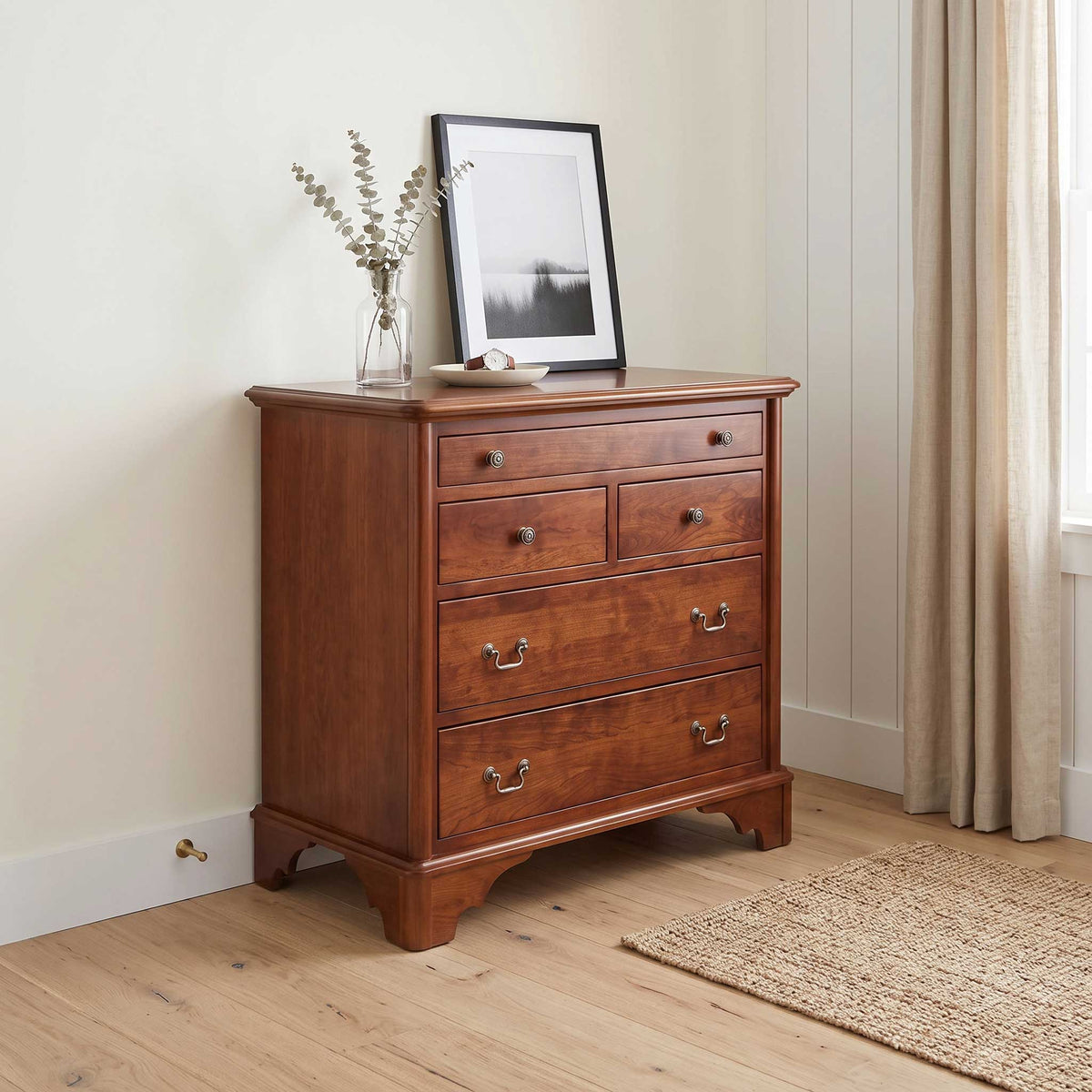 A warm brown wooden chest of drawers with six drawers and brass handles, styled in a bright room with a vase and framed print on top.