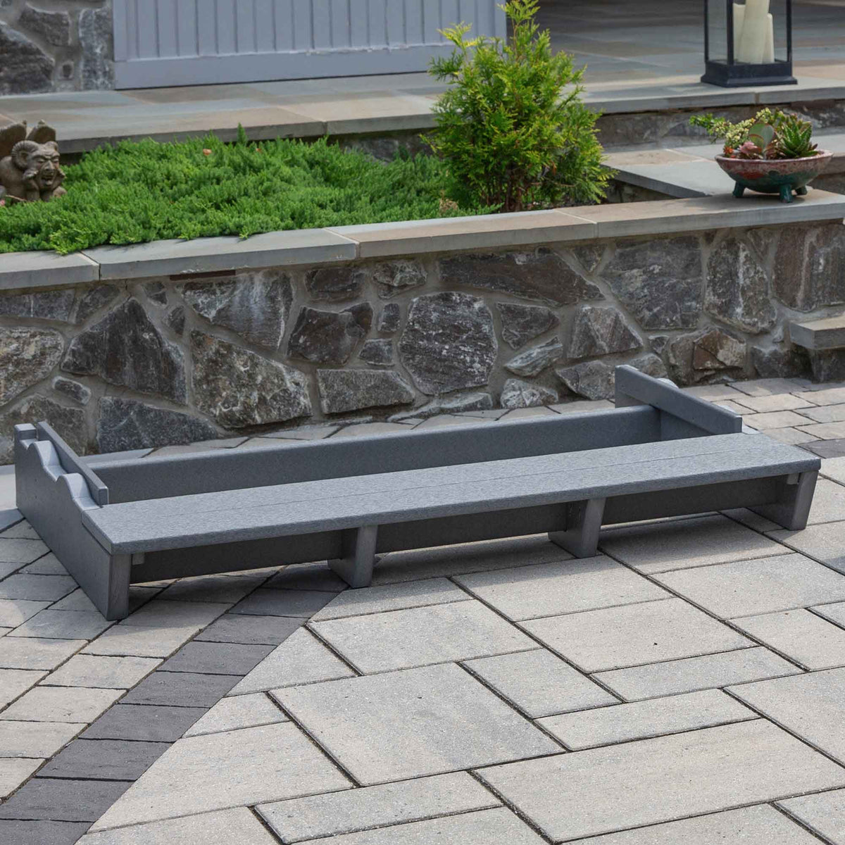 Dark gray poly lumber glider riser platform on a paver patio in front of a stone retaining wall and landscaped beds