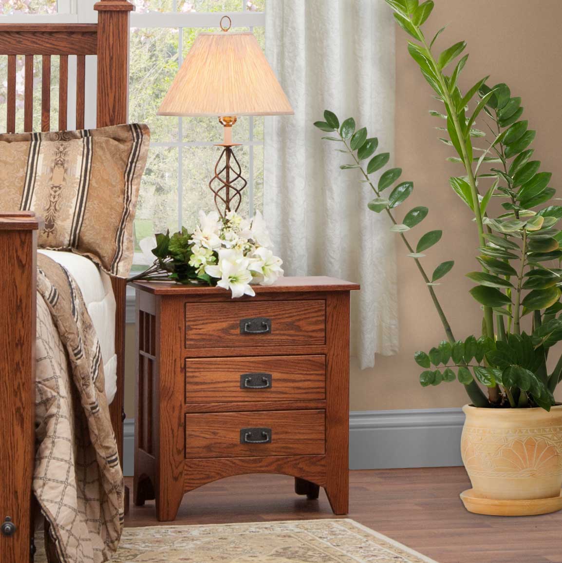 Mission oak three-drawer nightstand with black bail pulls beside a slatted bed, topped with a lamp and lilies