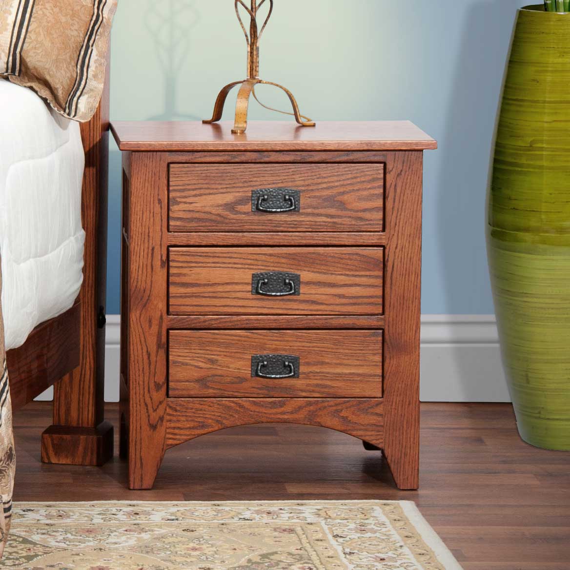 Mission oak three-drawer nightstand with black bail pulls beside a slatted bed, topped with a lamp and lilies