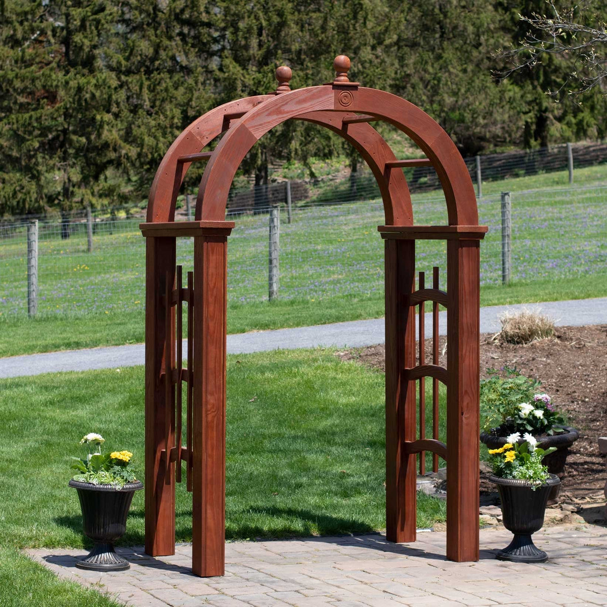 Cinnamon stained wood garden arbor with rounded arch top, ball finials and slat trellis sides on a paver walk