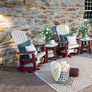 Pair of weatherwood and cherry Adirondack gliders with green pillows and round side tables on a stone patio