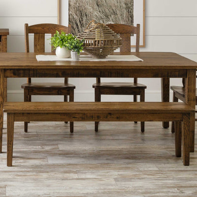 Rustic oak dining bench with a plank seat and tapered legs, tucked under a matching farmhouse table with greenery and a basket
