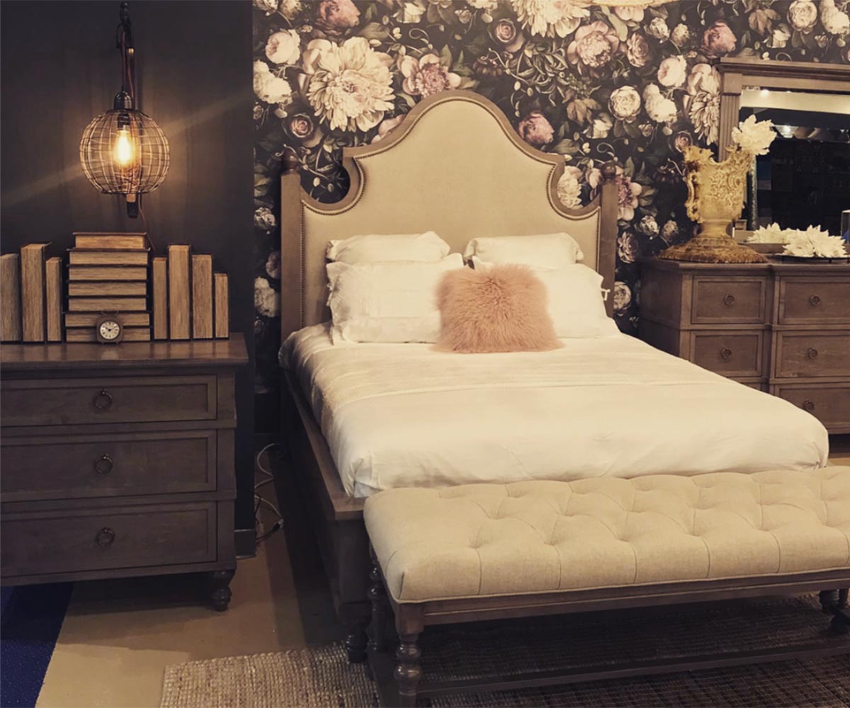 Upholstered bed with linen headboard staged against floral wallpaper with a tufted bench and nightstands