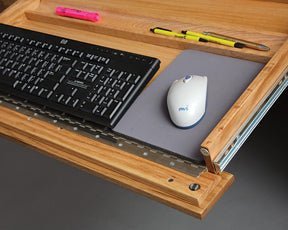 Pull-out keyboard tray of the oak desk holding a keyboard, mouse on a gray pad and pens on metal slides
