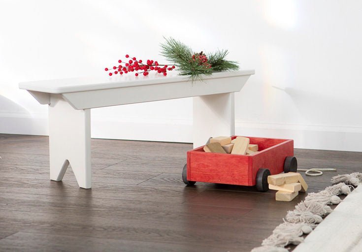 Red toy block wagon tipped with blocks spilling onto plank flooring under a white farmhouse bench with greenery