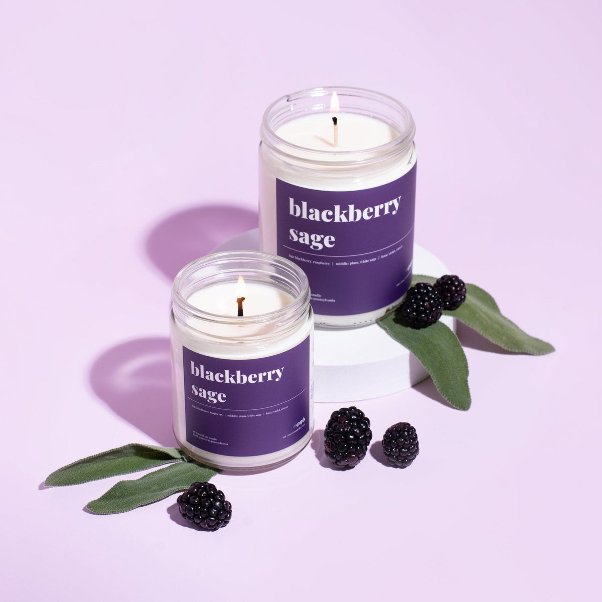 Two blackberry sage soy candles with deep purple labels burning beside fresh blackberries and sage leaves on lilac