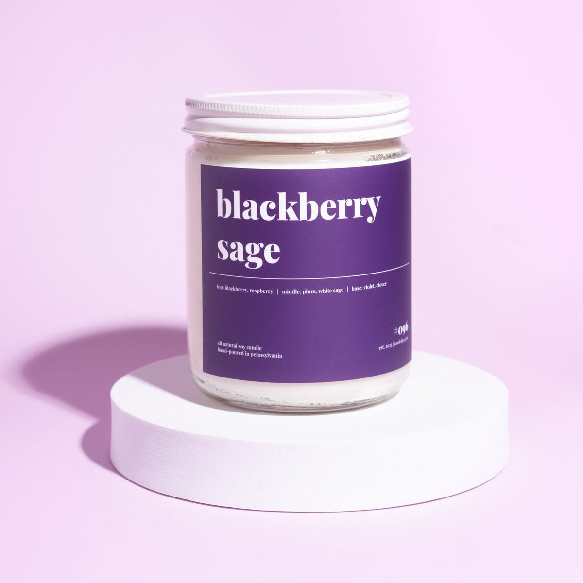 Standard blackberry sage soy candle with white lid and purple label displayed on a white riser against a lilac backdrop