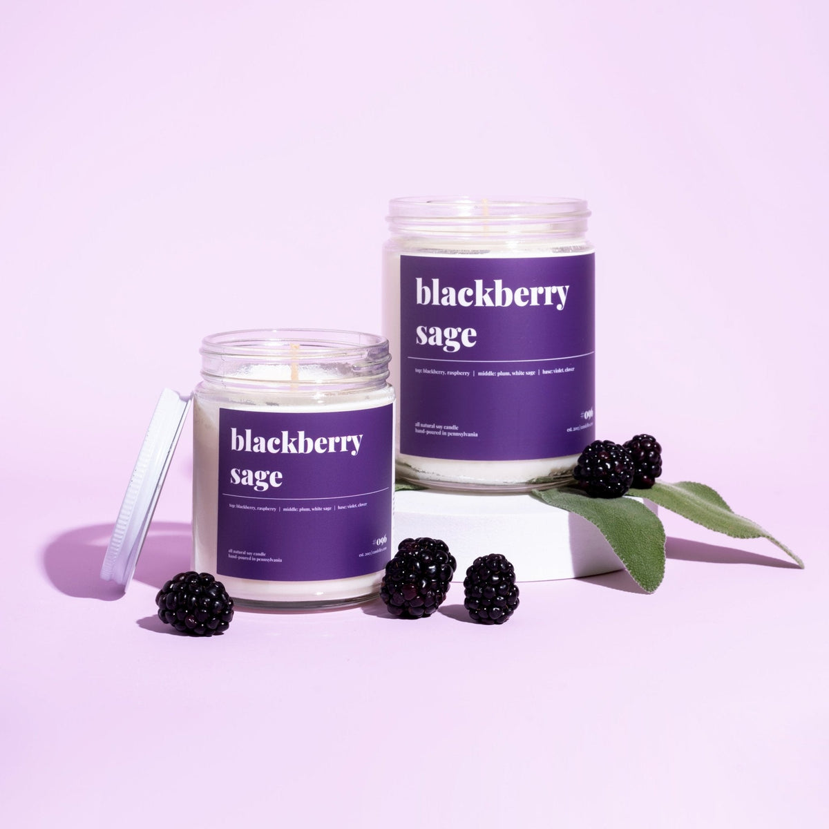 Two blackberry sage soy candles, one lidded and one lit on a white riser, with fresh blackberries and sage on lilac