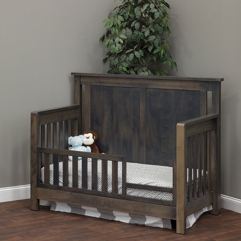 Gray panel crib converted to a toddler bed with a slatted safety rail and stuffed animals on chevron bedding
