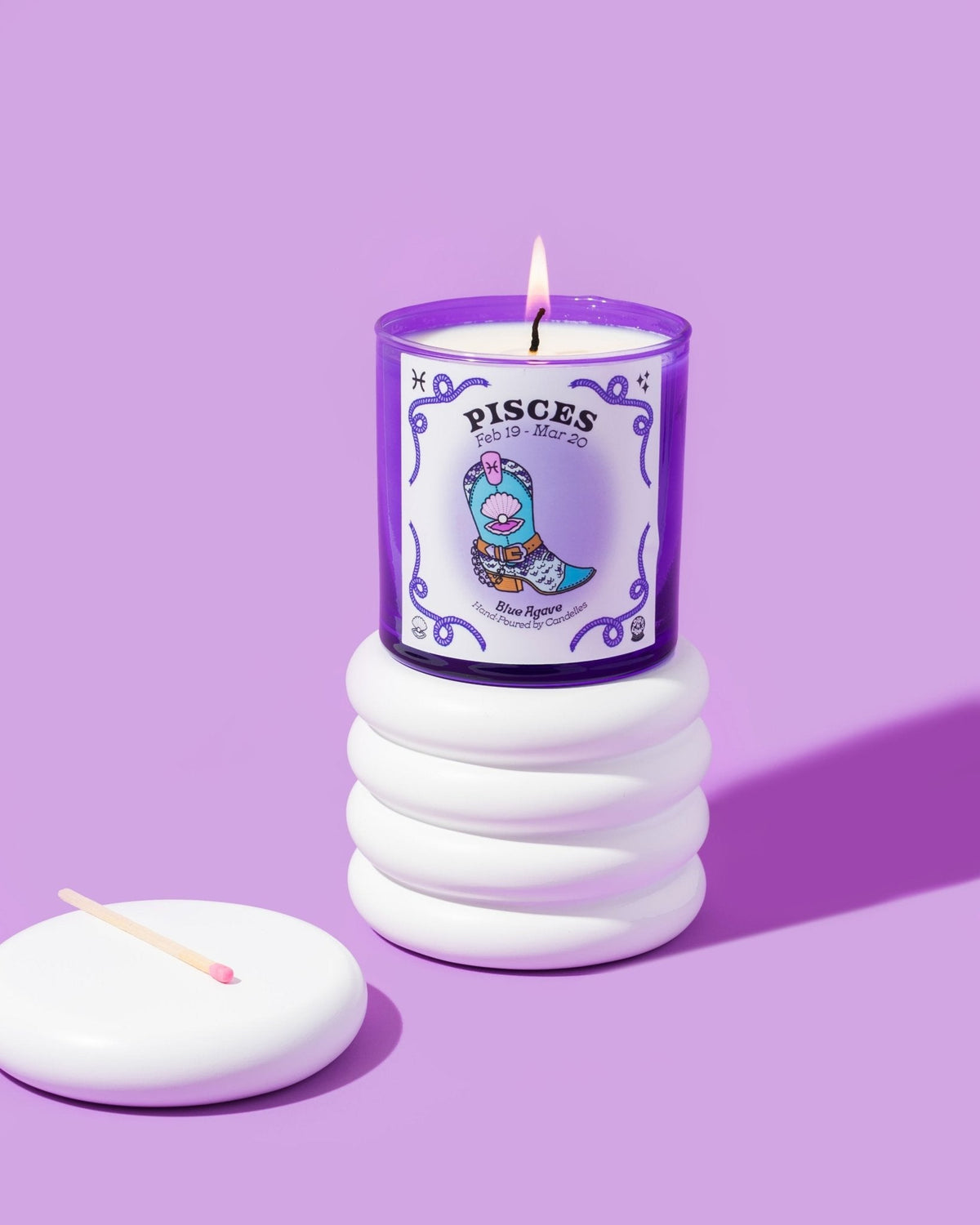 Blue Agave Pisces zodiac candle in a purple glass tumbler burning atop a white ribbed pedestal with a match and lid