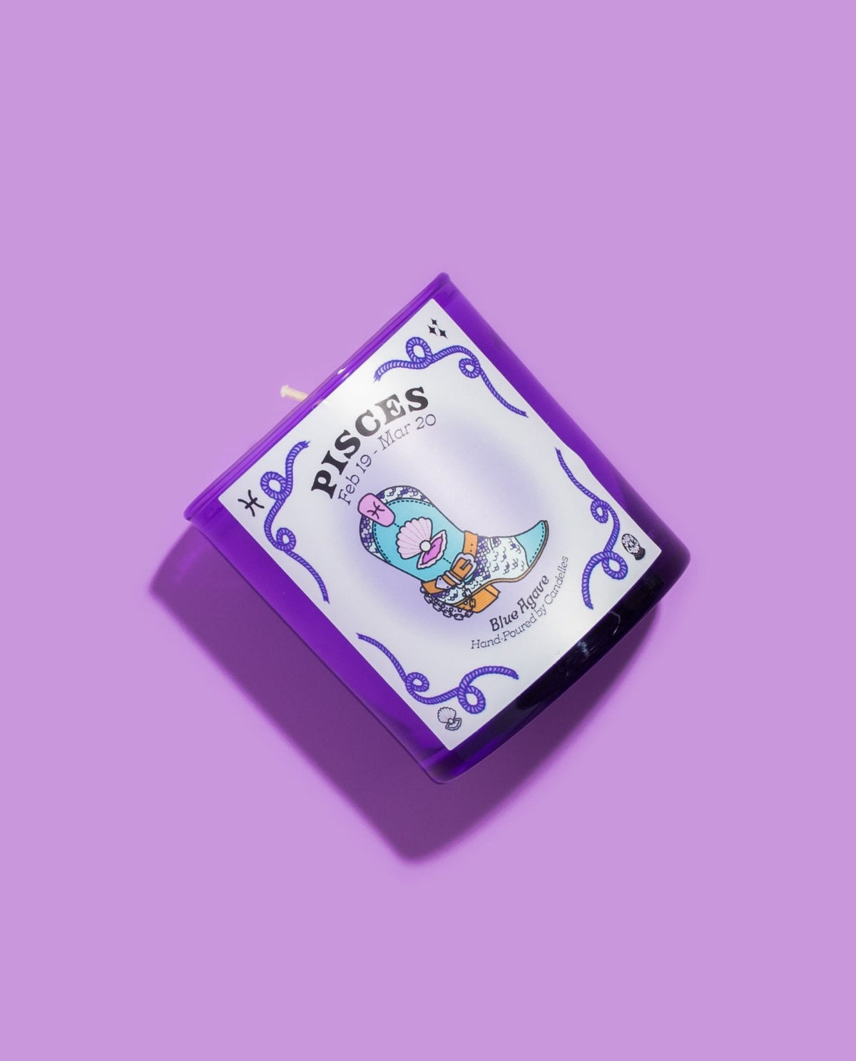 Purple glass Pisces zodiac candle tilted to show the illustrated cowboy boot label with blue agave scent on a lilac backdrop
