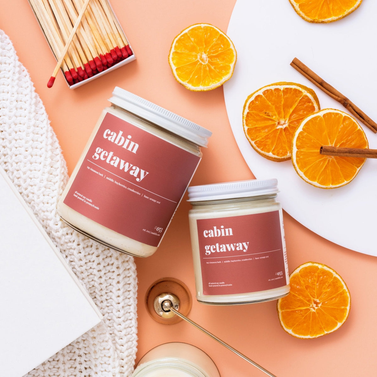 Overhead flat lay of two cabin getaway candles with dried orange slices, cinnamon sticks and matches on peach