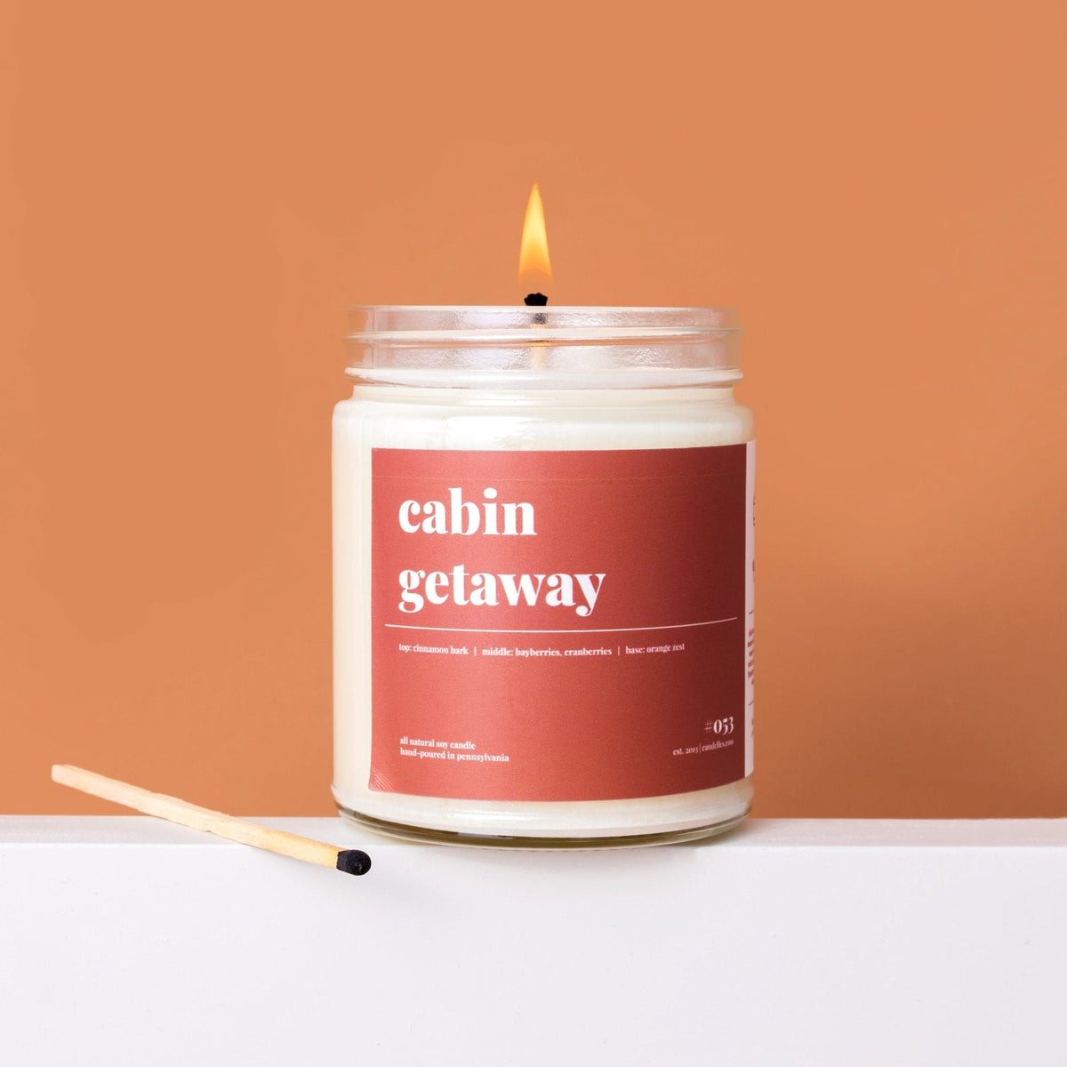Cabin getaway soy candle burning in a clear jar with a rust red label, spent match beside it on a terracotta backdrop