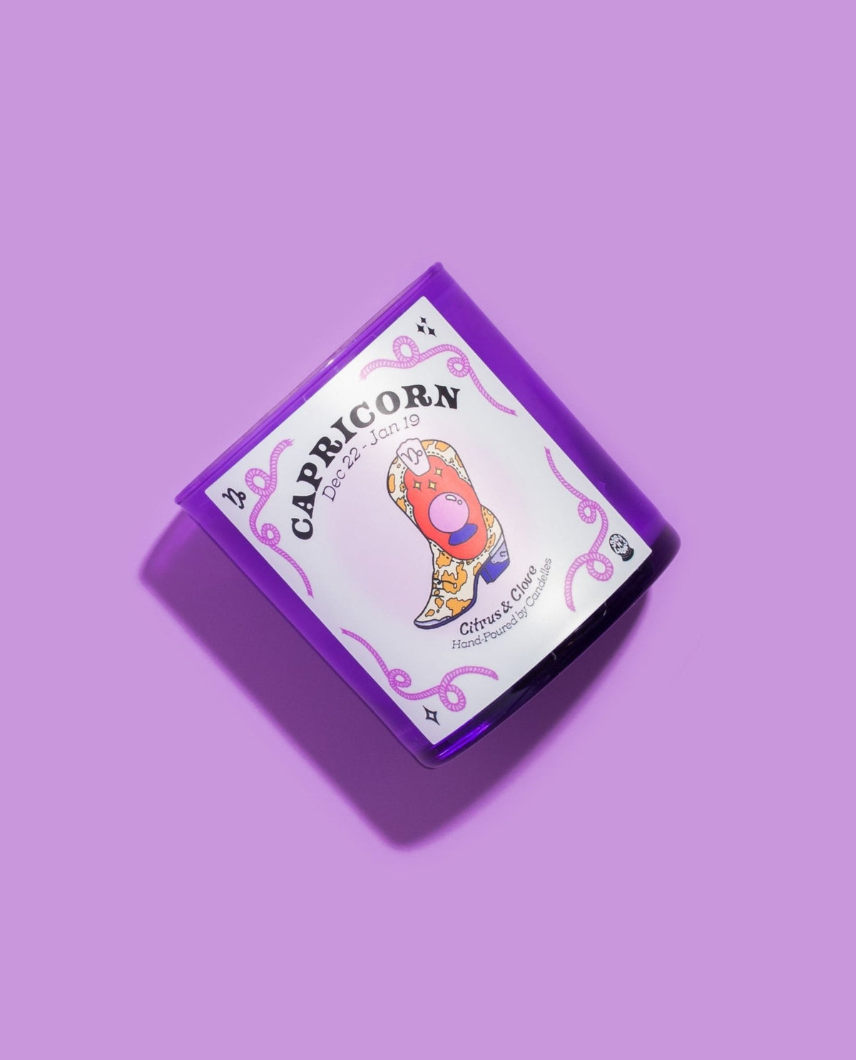 Capricorn citrus and clove candle tilted on a lilac background showing the illustrated pink cowboy boot label