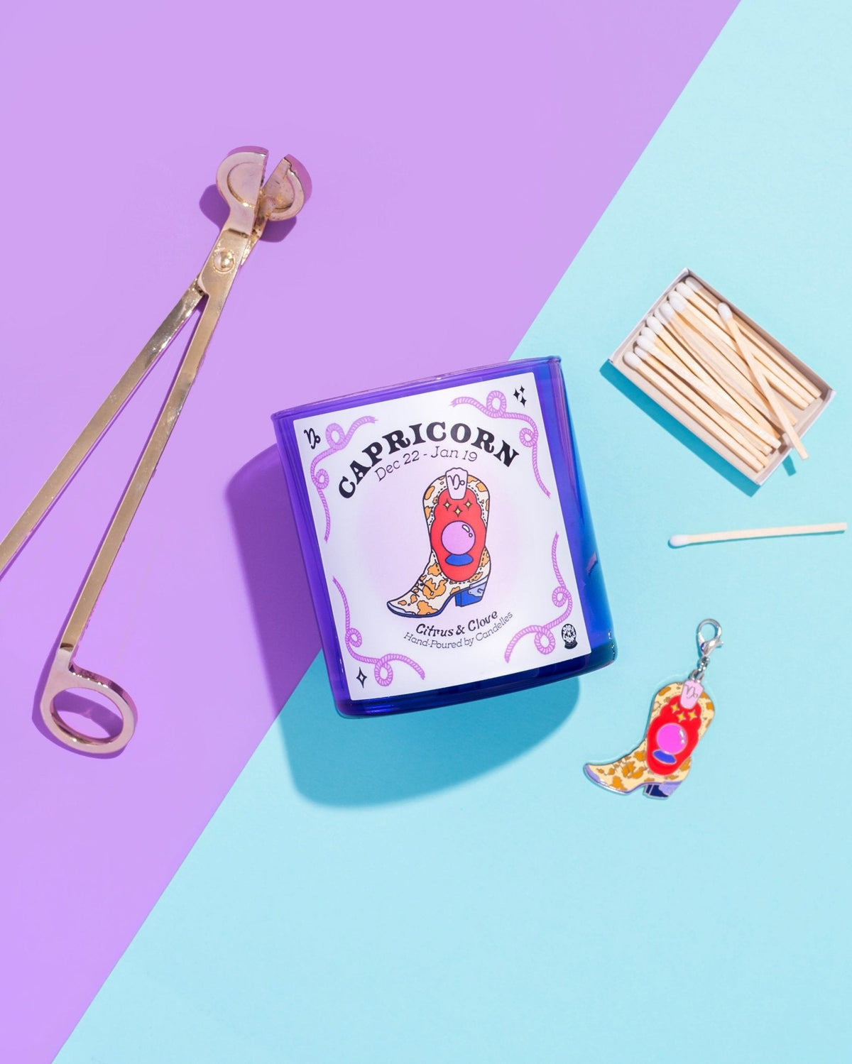 Citrus and clove Capricorn candle in a blue glass tumbler with wick trimmer, matches and a boot charm on purple and blue